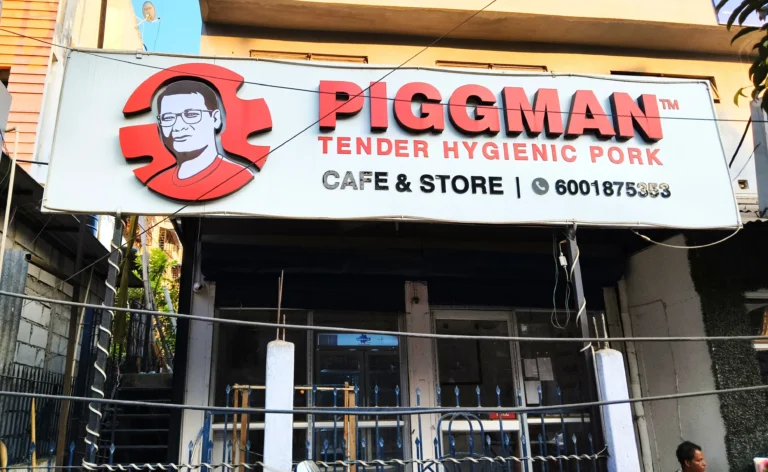 Front facade of a Piggman outlet displaying the Piggman logo and signage reading ‘Piggman Tender Hygienic Pork – Cafe & Store’, with the storefront and surrounding street visible.